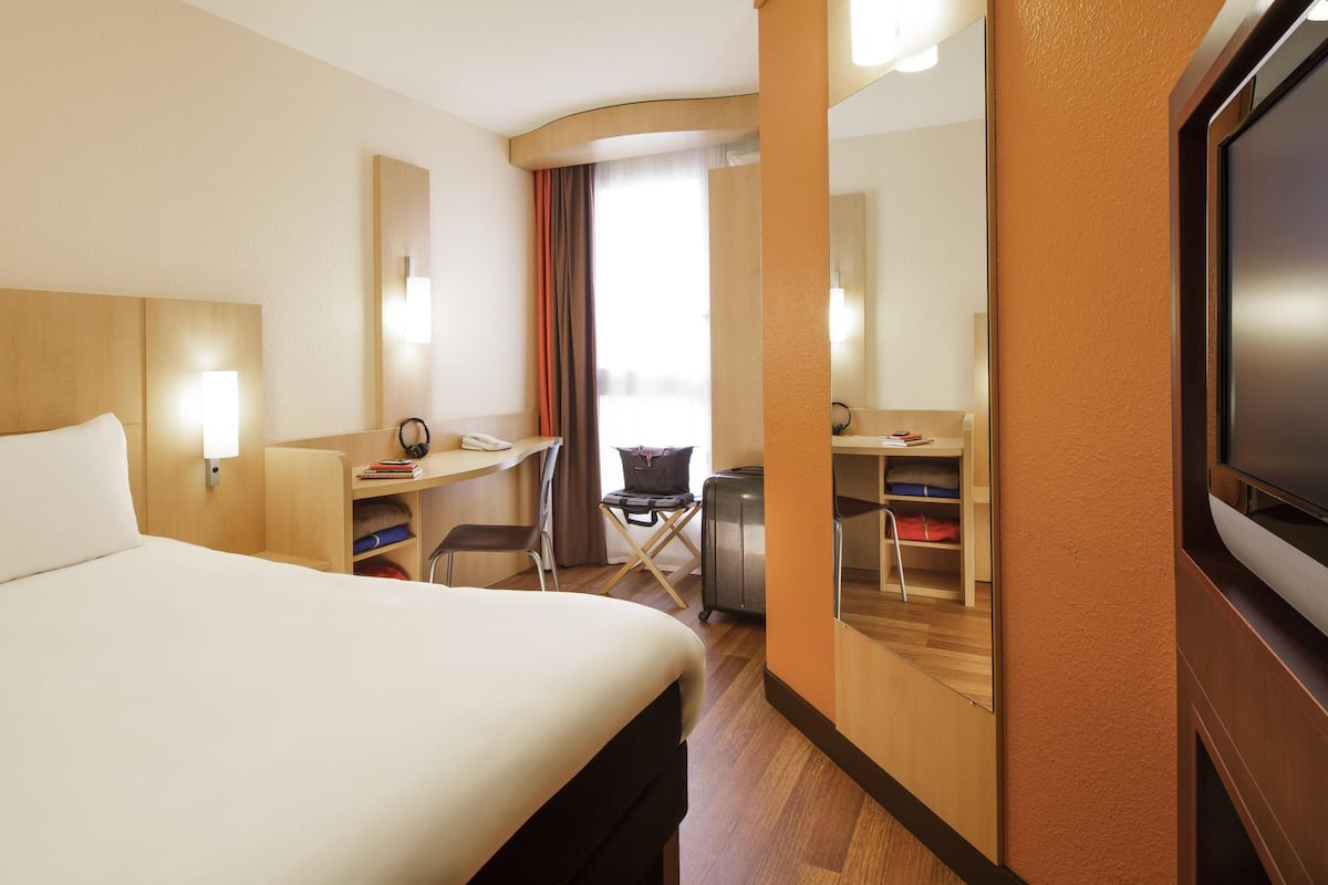 A hotel room with a bed, desk, chair, TV, and a large mirror.