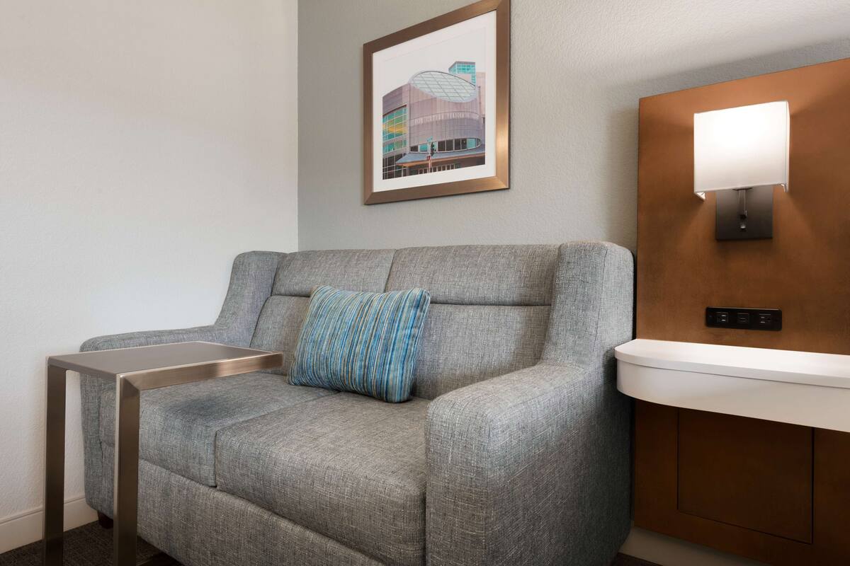 A modern hotel room with a grey sofa, a striped pillow, a framed artwork of a building, and a wall-mounted light fixture.
