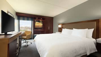 A hotel room with a large bed, a desk with a TV, a chair, and a window with a view of a cityscape.