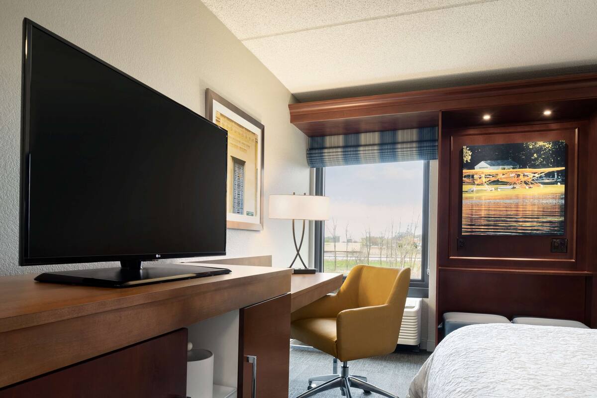 A hotel room with a large flat-screen TV, a wooden desk, a yellow chair, a bed, and a window with a view of a landscape.
