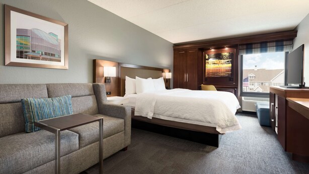 A hotel room with a bed, a sofa, a small table, a TV, and a window with a view of buildings.