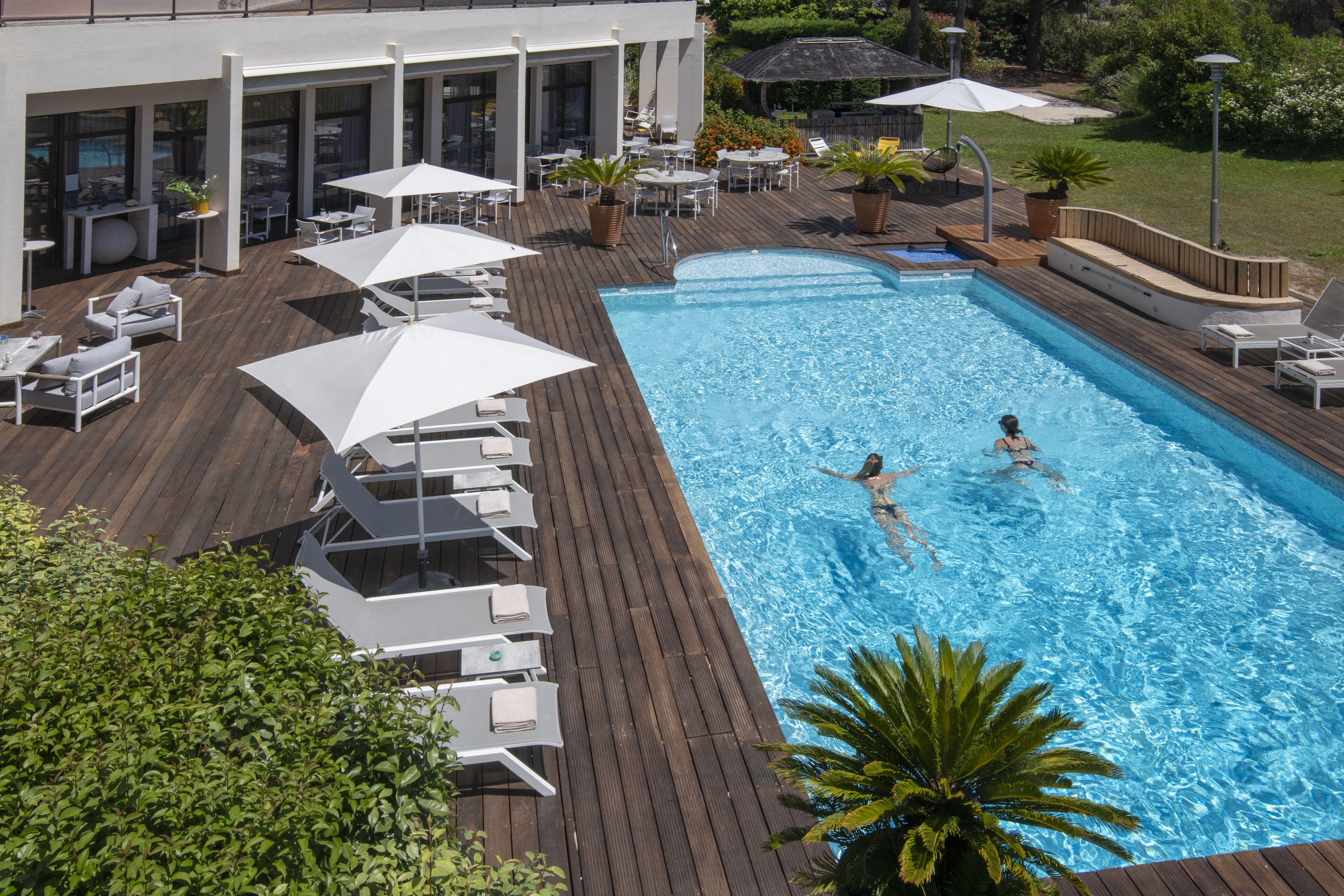 Ein rechteckiger Pool mit zwei Schwimmern, umgeben von weißen Liegestühlen und Sonnenschirmen, auf einem Holzboden mit Blick auf ein Gebäude und Grünflächen.
