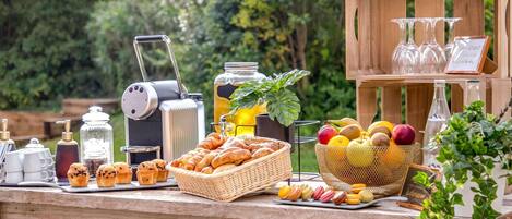 A wooden table with a portable coffee maker, a jar of honey, a basket of croissants, a bowl of fruit, and a wooden crate with glasses and a sign.
