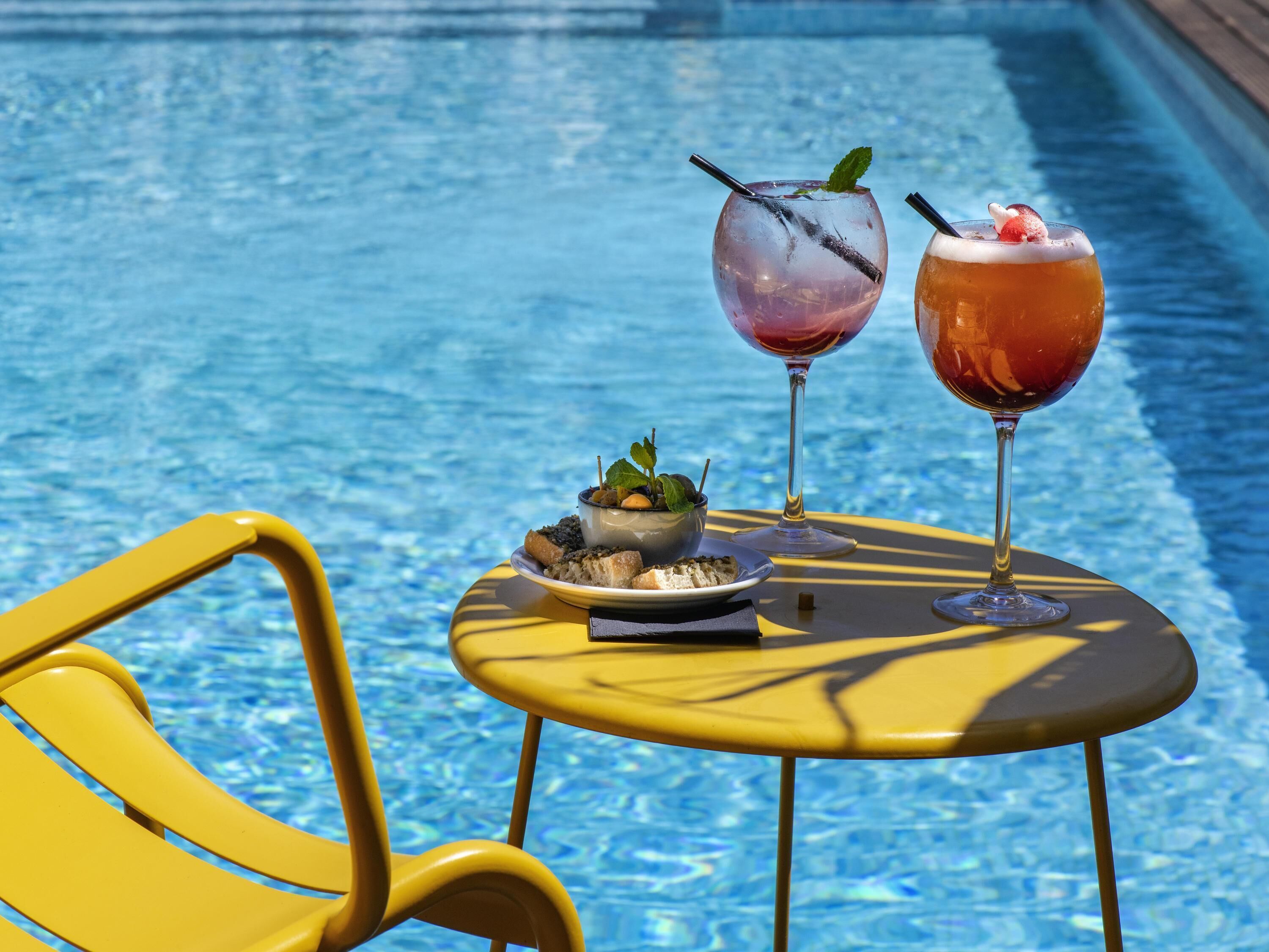 Mesa junto a la piscina con dos bebidas, un bol de aceitunas y una bandeja de pan.