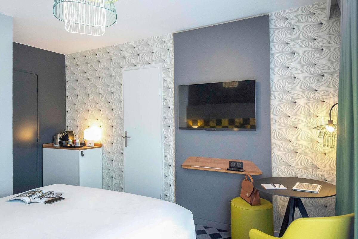 Habitación de hotel con cama, mesita de noche, lámpara, silla y televisor montado en la pared.