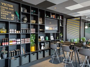 Un interior moderno con un letrero de cafetería, estantes con diversos artículos y una zona de estar con taburetes de bar.