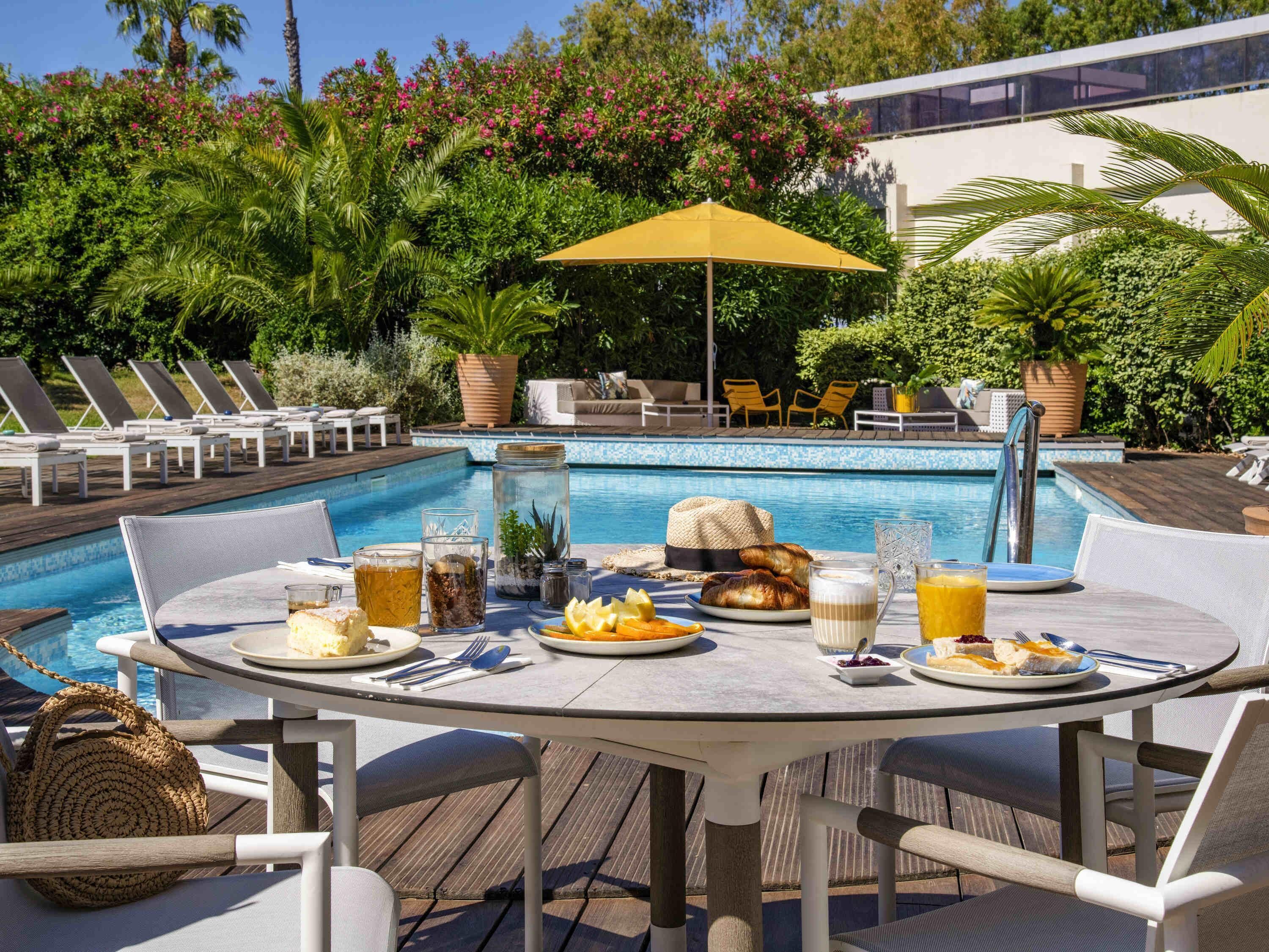 Un área de comedor junto a la piscina con una mesa dispuesta para dos, rodeada de sillones, una piscina y abundante vegetación.