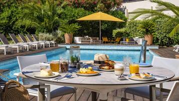 Un área de comedor junto a la piscina con una mesa dispuesta para dos, rodeada de sillones, una piscina y abundante vegetación.