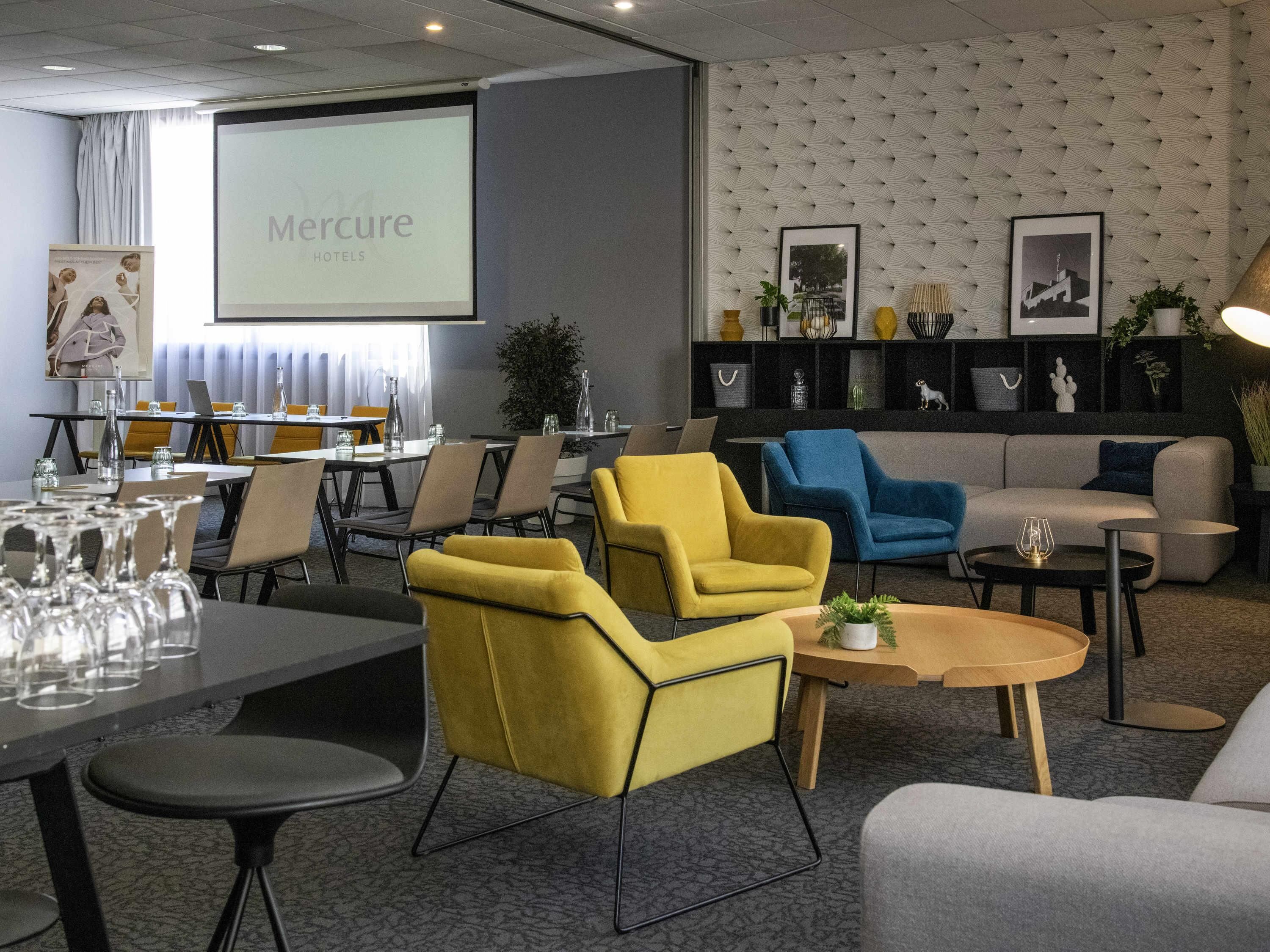 Une salle de conférence équipée d’un écran de présentation, de chaises jaunes et bleues, d’un canapé et d’un espace d’accueil agrémenté de tableaux et de plantes.