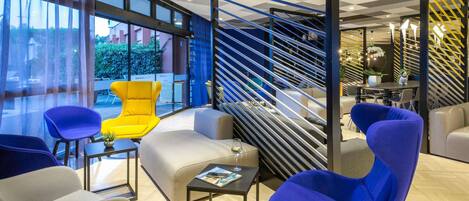 A modern hotel lobby with a checkered ceiling, a staircase with blue and yellow railings, and a seating area with colorful chairs.