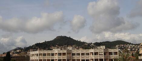 A multi-story building with a symmetrical facade, balconies, and a view of a hill with scattered houses.