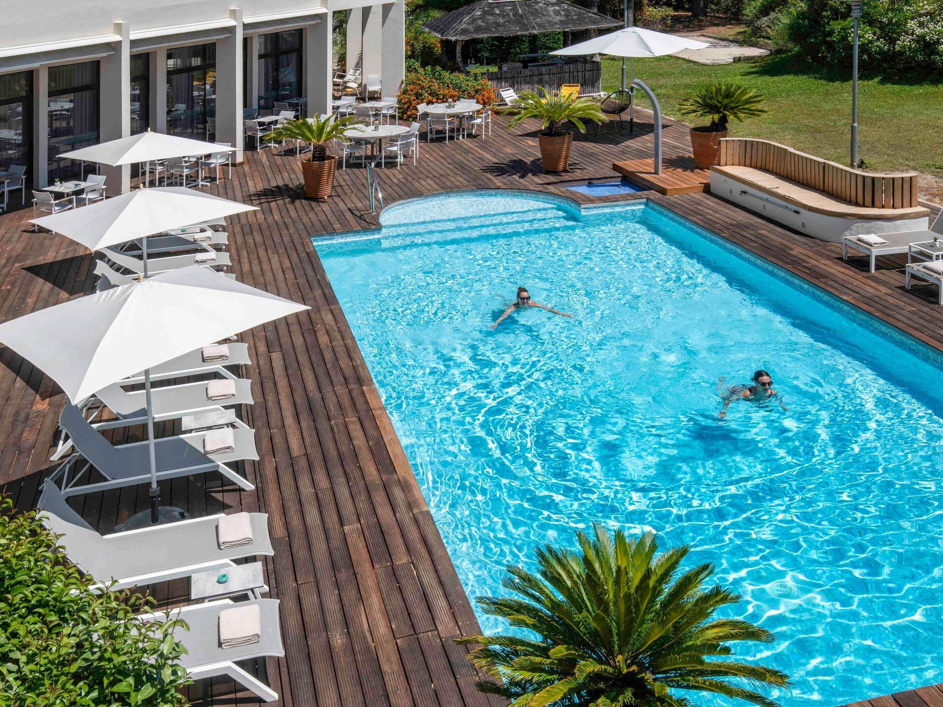 Una piscina con sillas de descanso y sombrillas, rodeada por una terraza de madera y plantas en macetas.