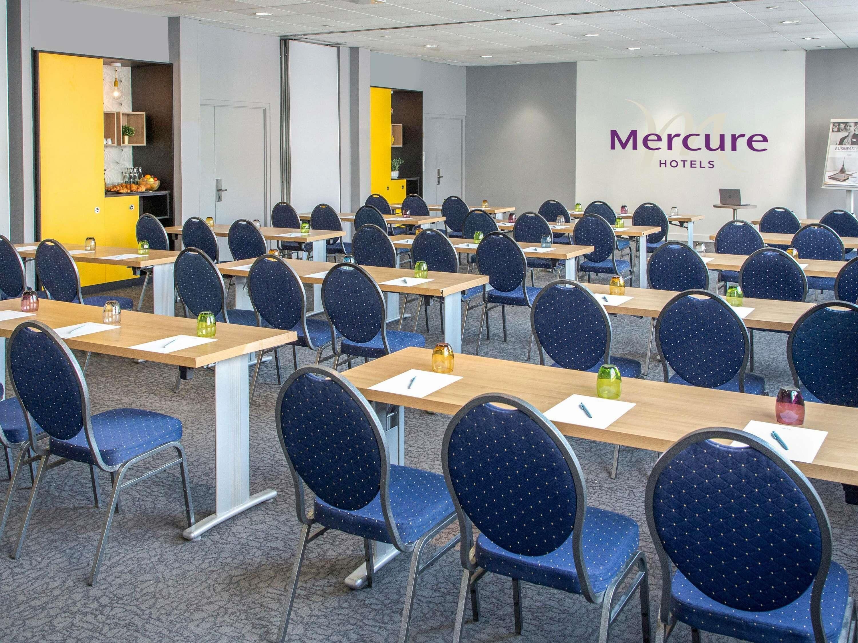 Uma sala de conferências com fileiras de mesas e cadeiras, uma divisória amarela e o letreiro 'Mercure Hotels'.