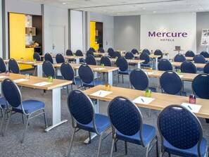 Een congreszaal met rijen tafels en stoelen, een geel scherm en een 'Mercure Hotels' bord.