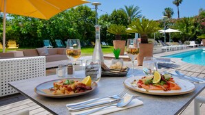 A dining table with two plates of food, a bottle, and glasses of water, set outdoors with a pool and lounge chairs in the background.
