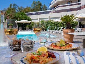 Une table à manger avec des assiettes remplies de nourriture, des verres de vin, et une piscine en arrière-plan.