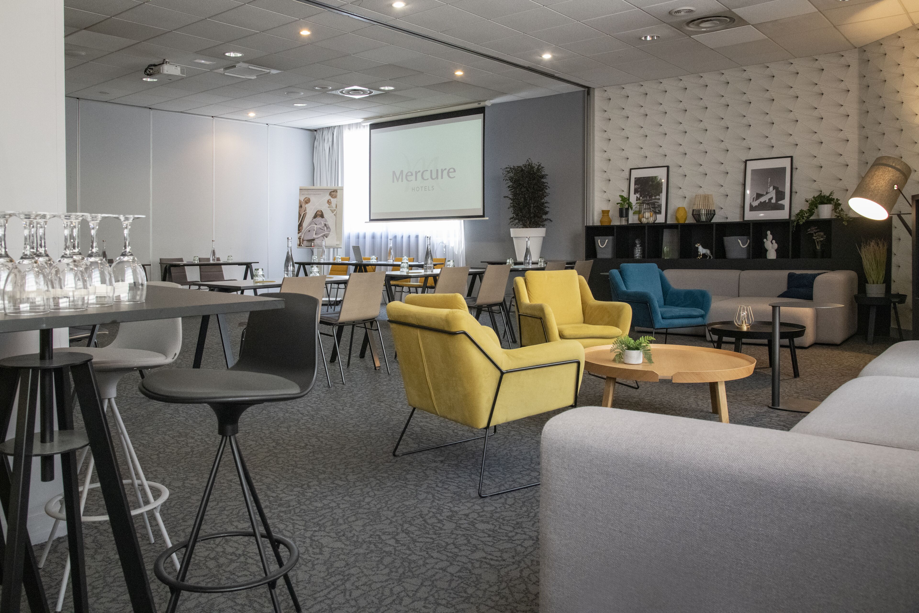 A modern conference room with a presentation screen, yellow and blue chairs, a grey sofa, and a bar area with stools.