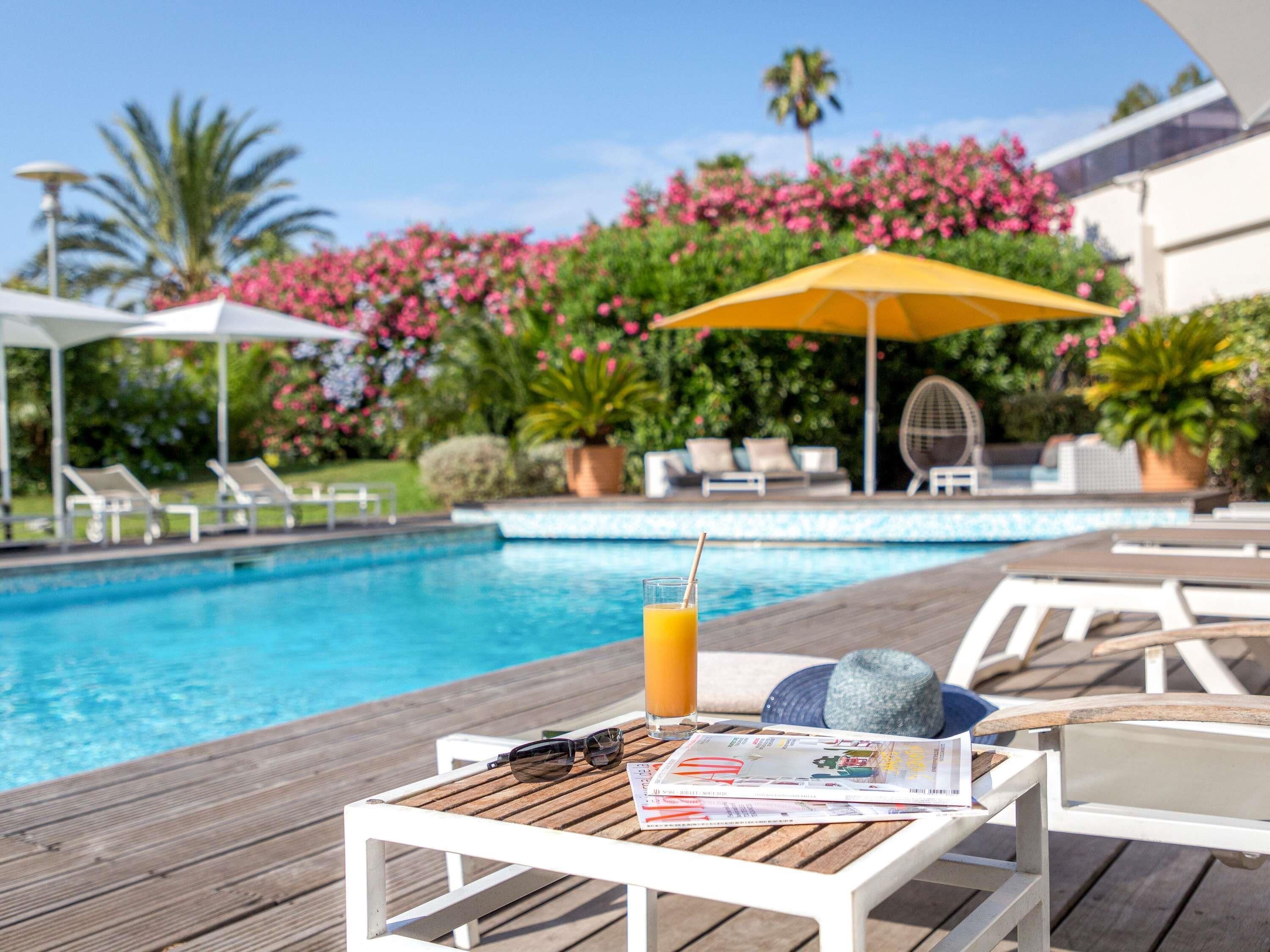 A poolside area with lounge chairs, a wooden table with a magazine, a drink, and a sun umbrella.