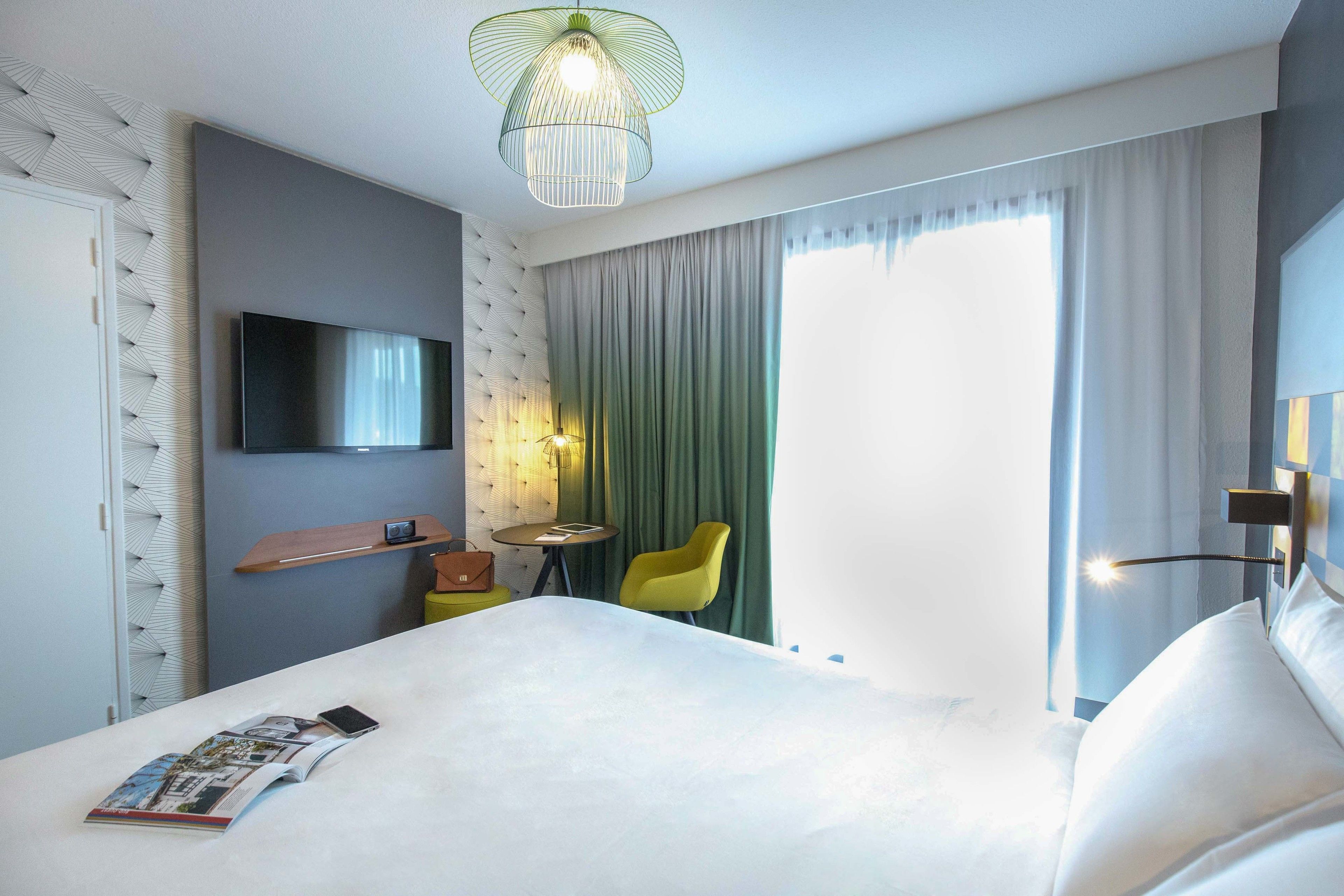 Een hotelkamer met een groot bed, een flatscreen televisie aan de muur, een klein tafeltje met een stoel en een raam met gordijnen.