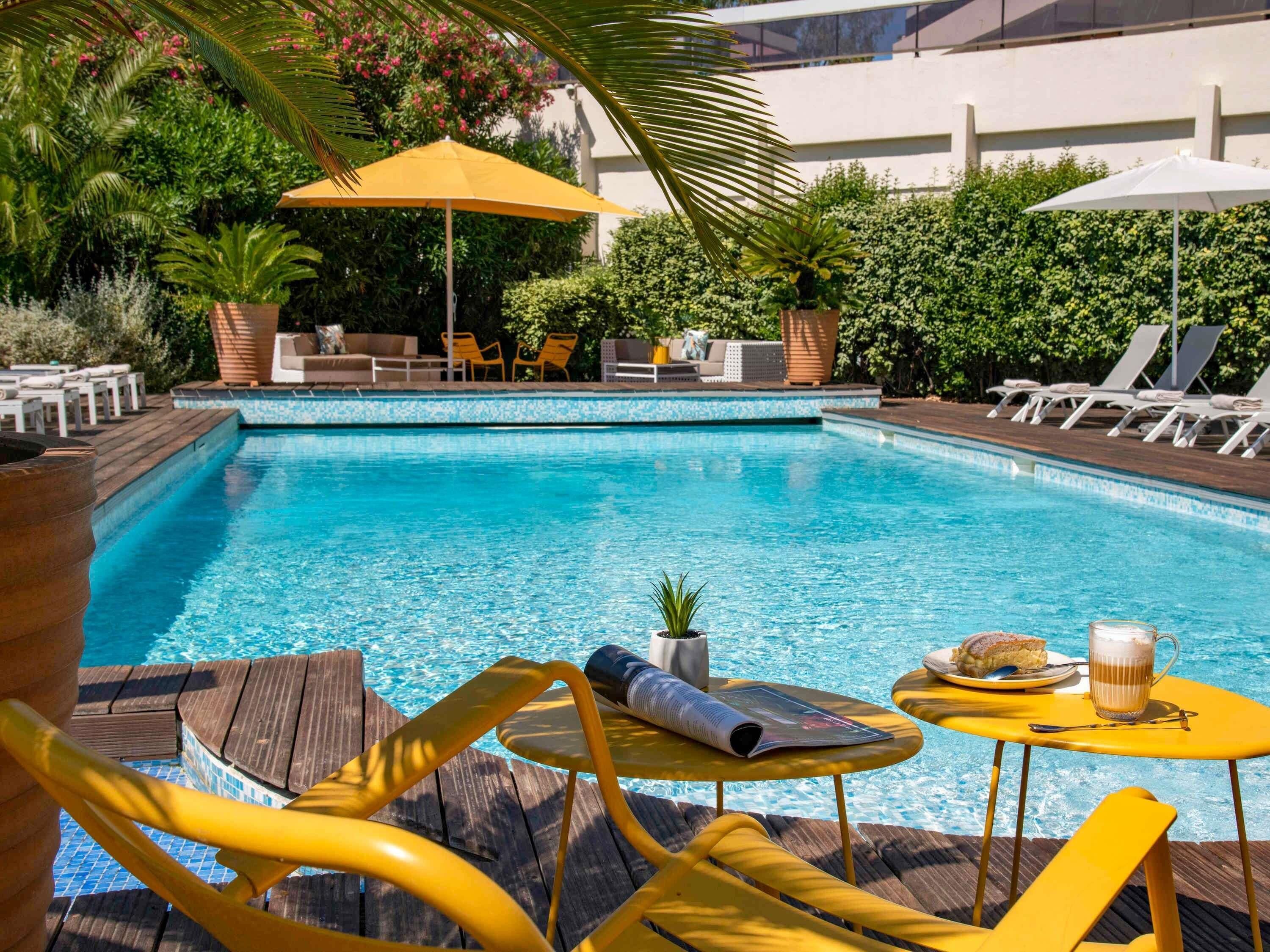 A swimming pool with lounge chairs and umbrellas, surrounded by greenery and a tiled deck.