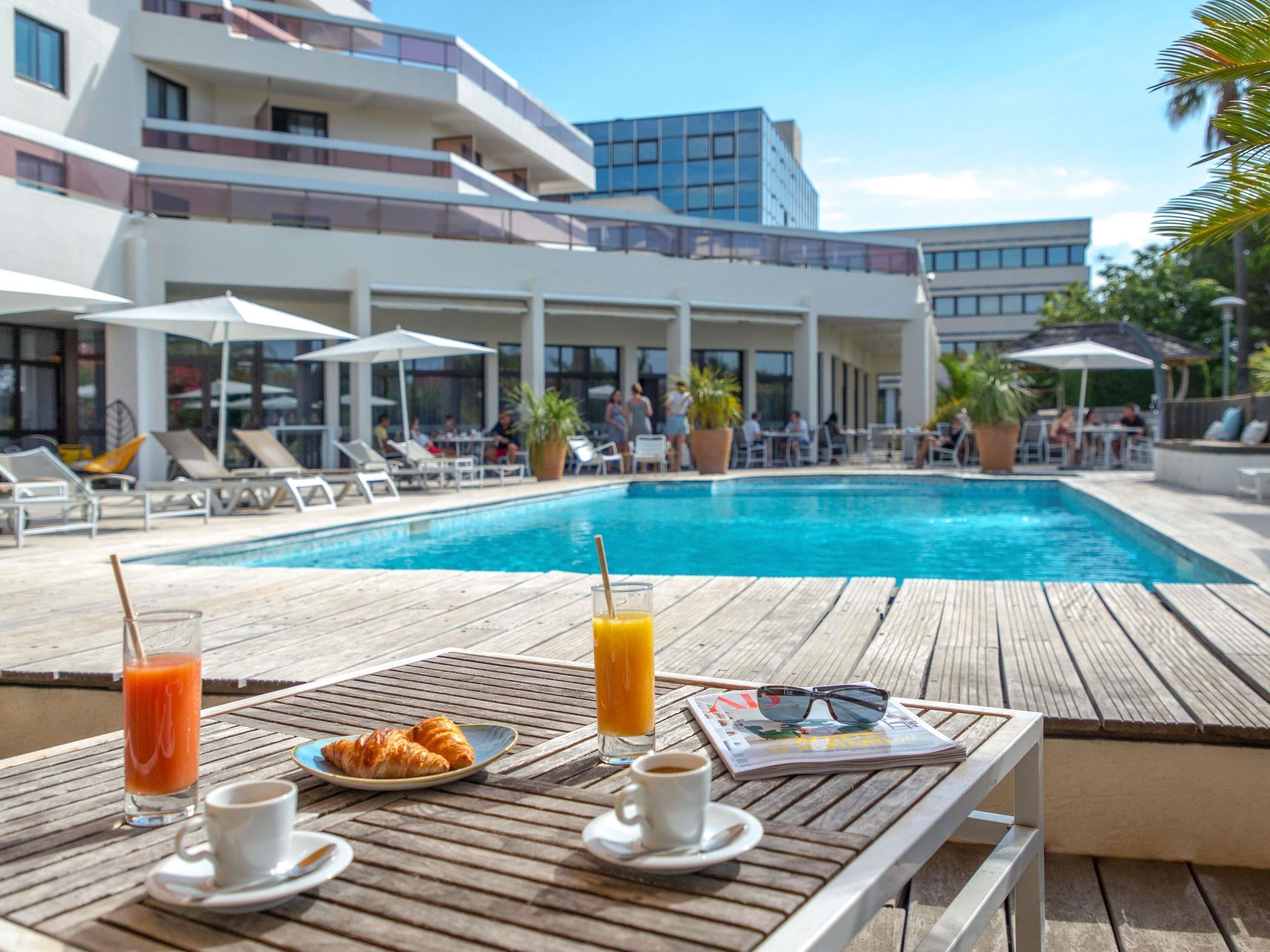 A poolside area with lounge chairs, a wooden table with breakfast items, and a view of a modern building in the background.