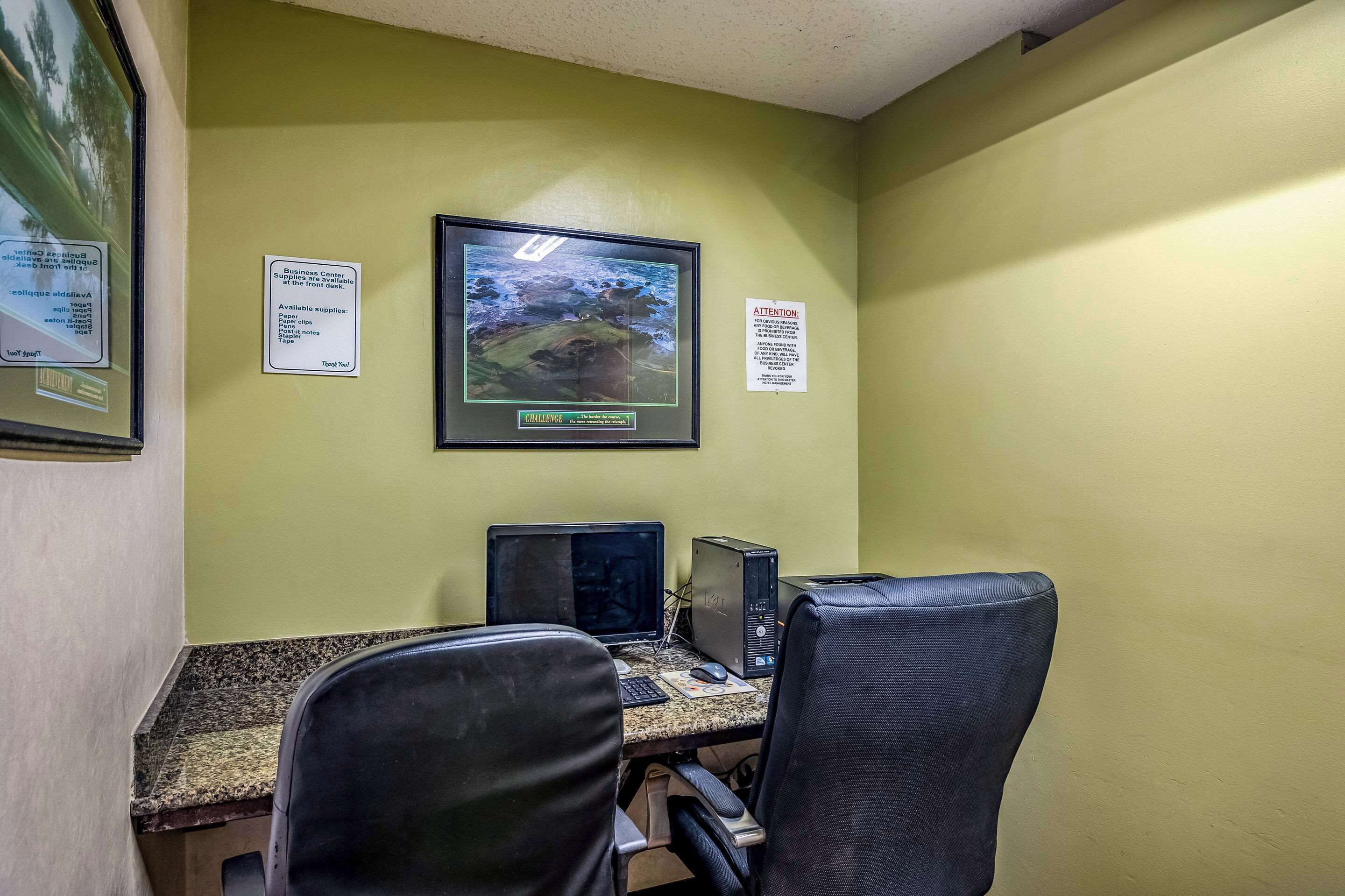 A business center with a desktop computer, two monitors, and two office chairs. There are signs on the wall indicating available supplies and a business center location.
