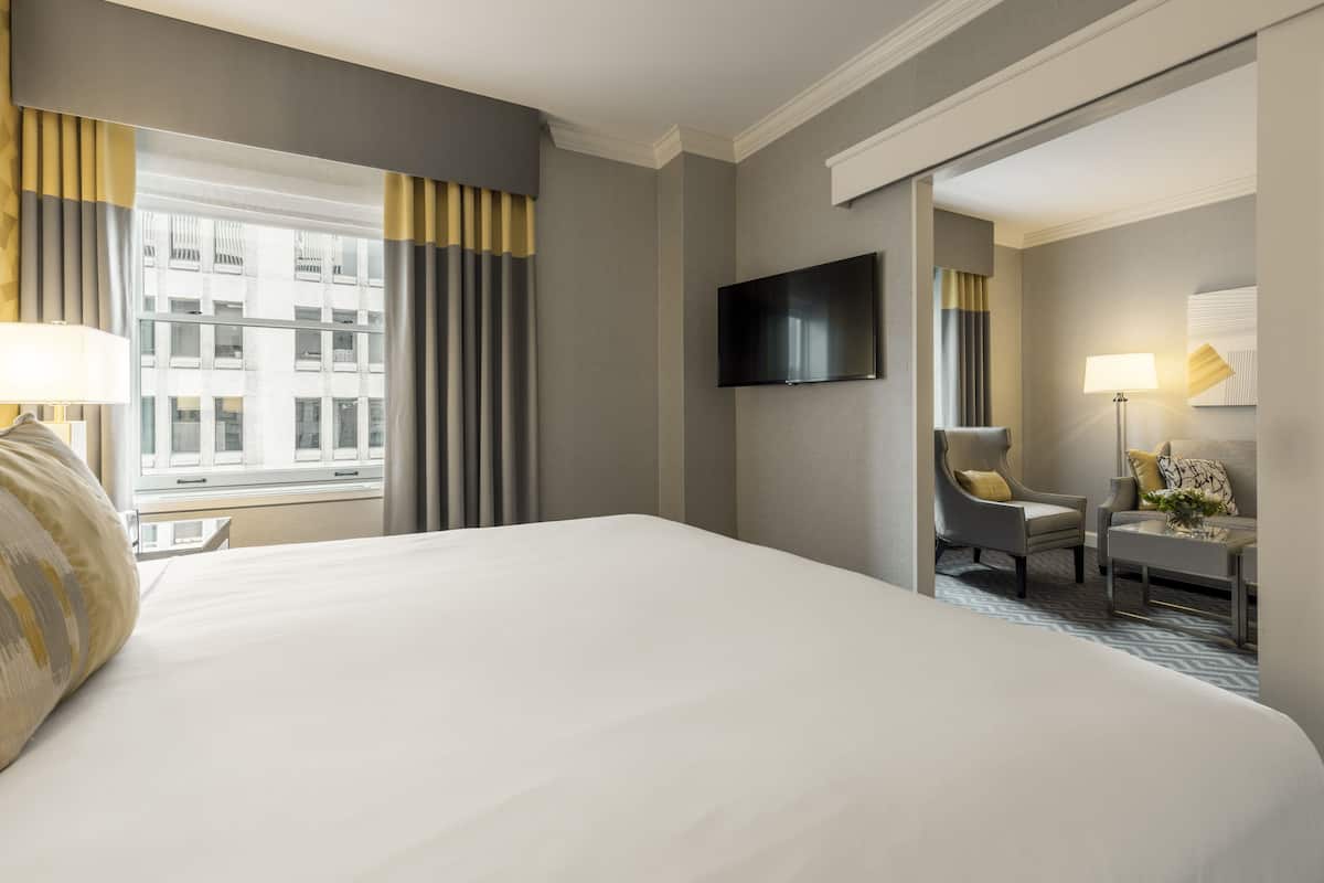 A hotel room with a large bed, a television mounted on the wall, a chair, a small table, and a window with curtains.