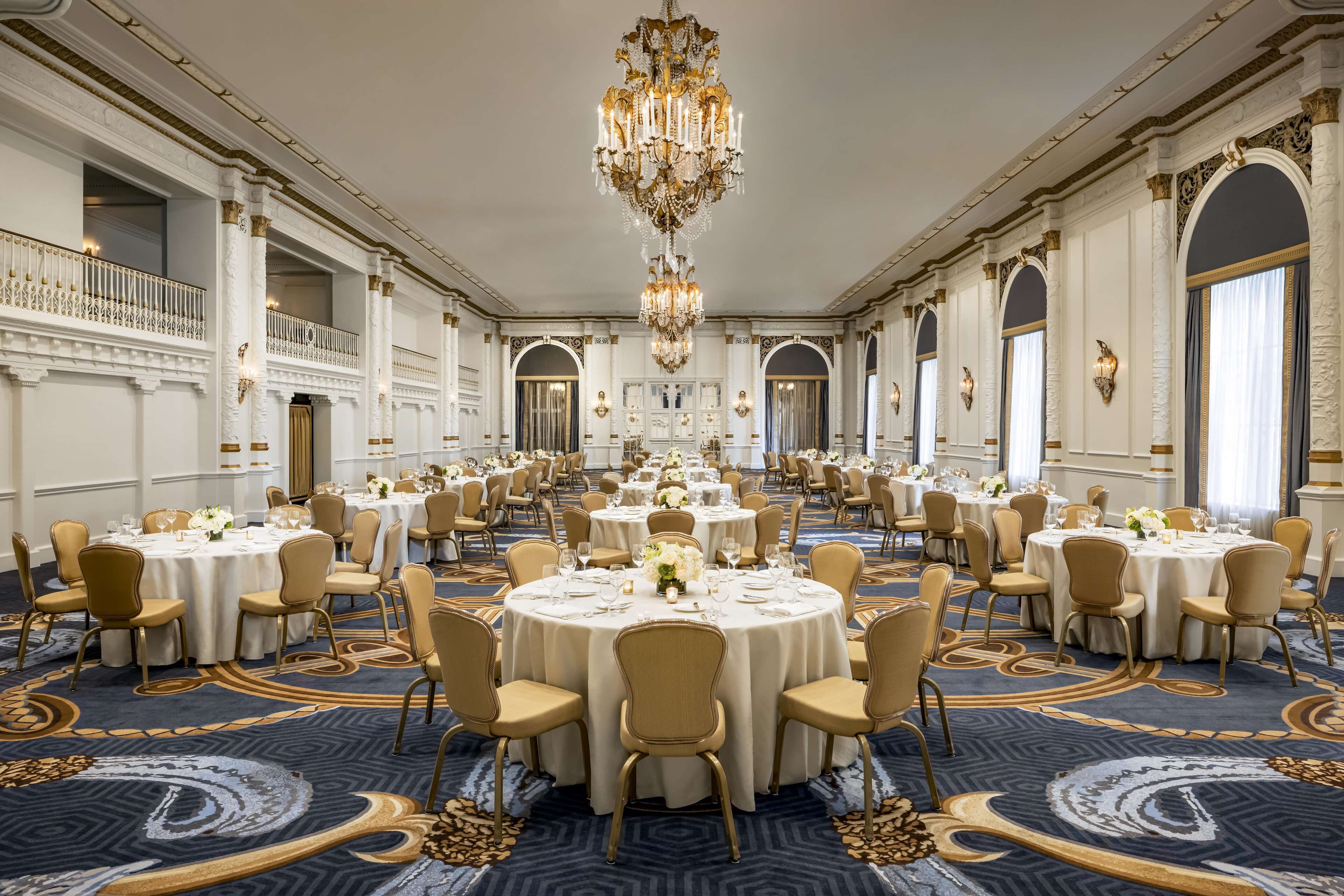 A grand banquet hall with multiple round tables, ornate chandeliers, and classic architectural details.