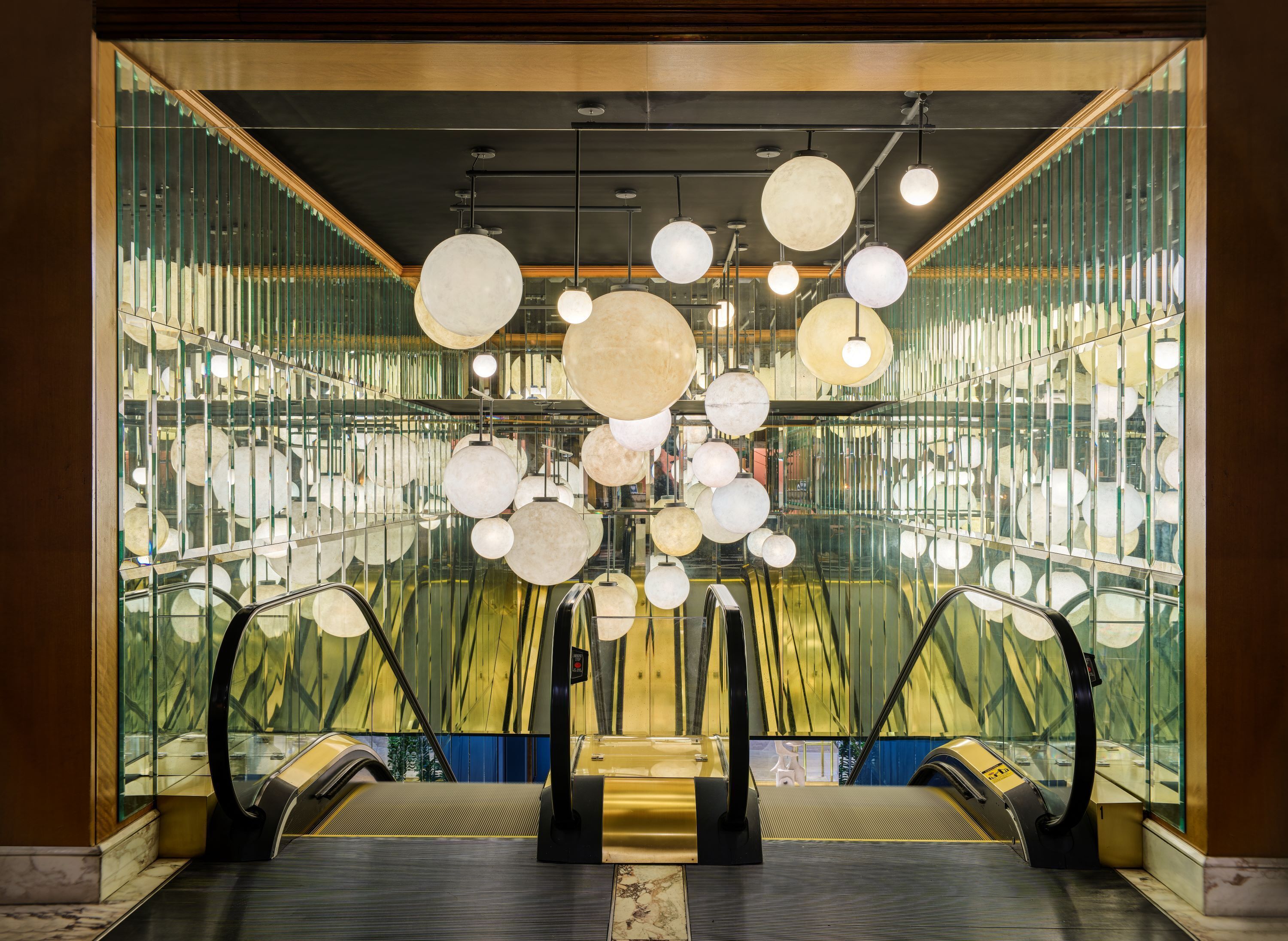 An interior with glass partitions, multiple escalators, and hanging spherical light fixtures.