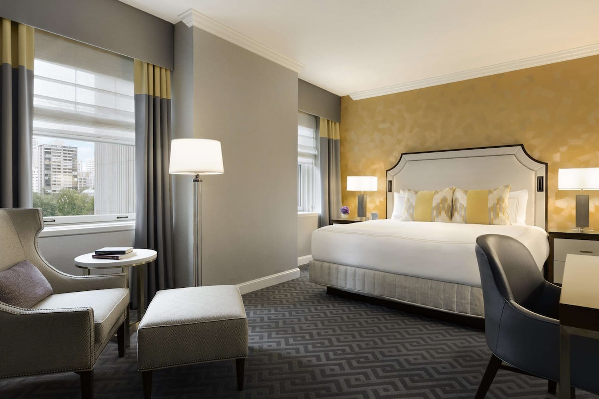A hotel room with a large bed, two armchairs, a small round table, and a view of the city through the window.