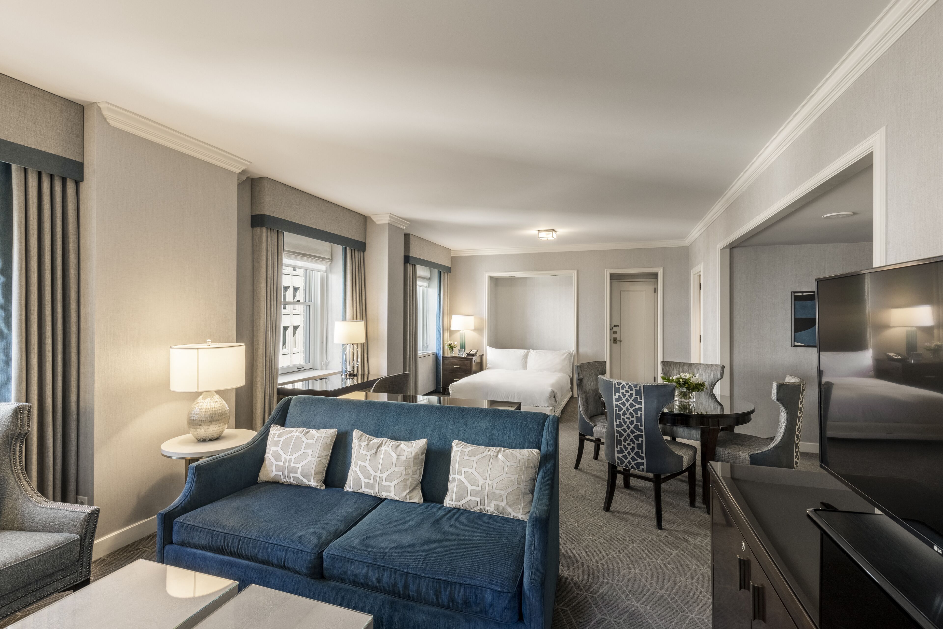 Ein Hotelzimmer mit blauem Sofa, einem Esstisch mit Stühlen, einem Fernseher, einem Bett und einem kleinen Schreibtisch mit Lampe.