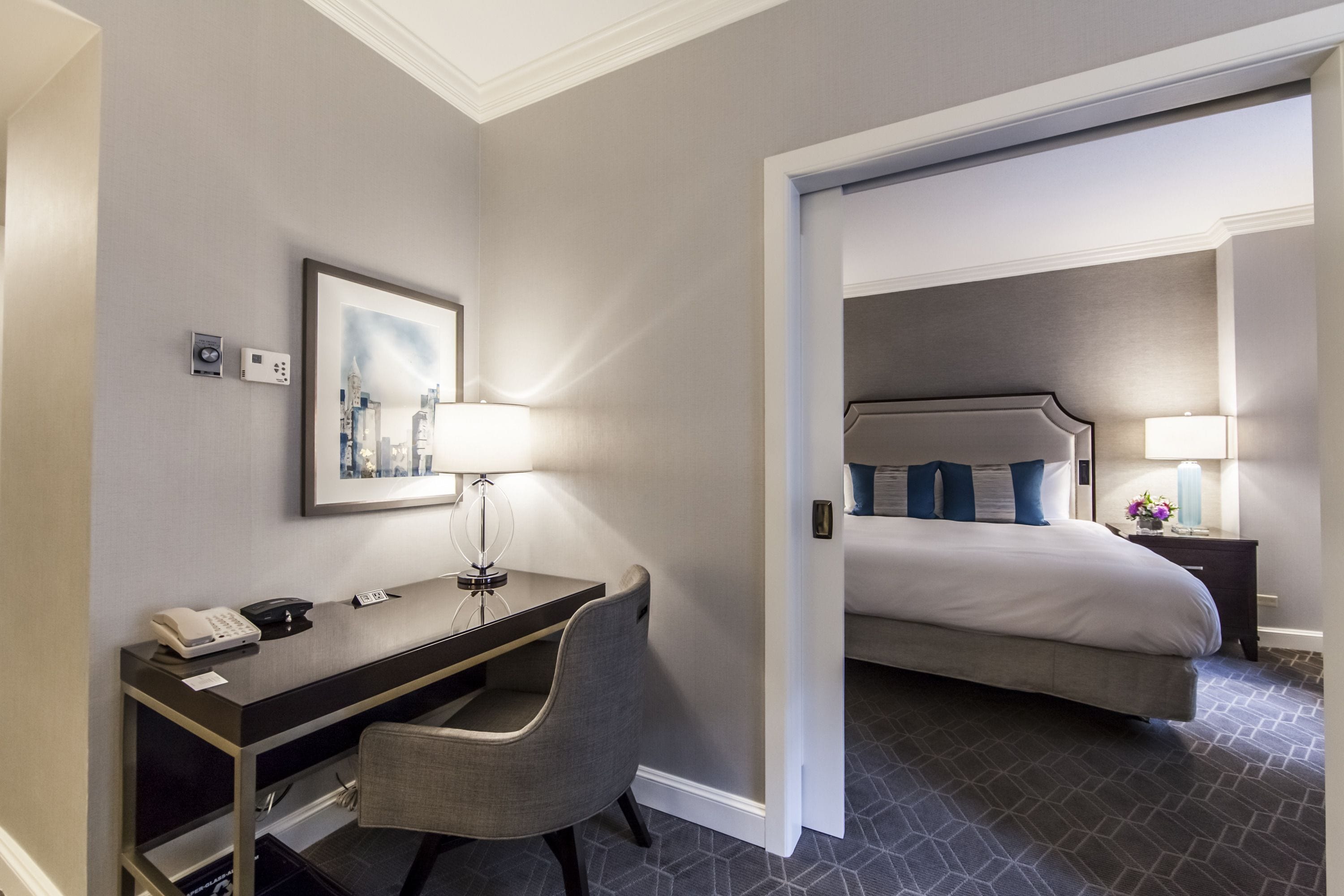 A hotel room with a desk, chair, bed, and a painting on the wall.