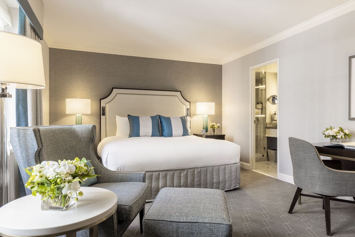 A hotel room with a large bed, two bedside tables with lamps, a round table with a vase of flowers, a grey armchair, and a desk with a chair.