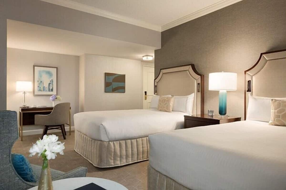 A hotel room with two beds, a desk, a chair, and a nightstand with a lamp.