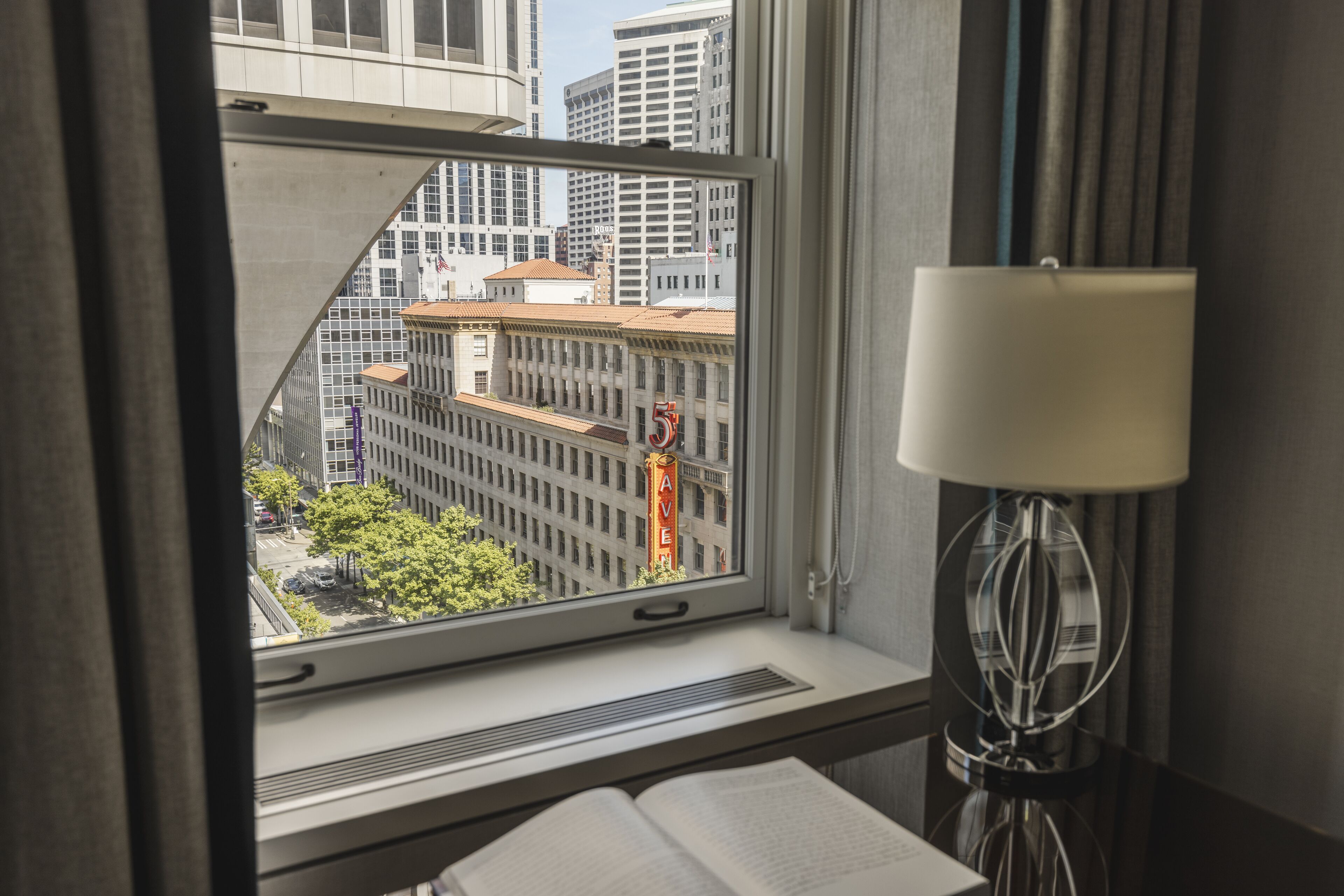 A hotel room with a window view of a cityscape, a lamp, and a book on the windowsill.