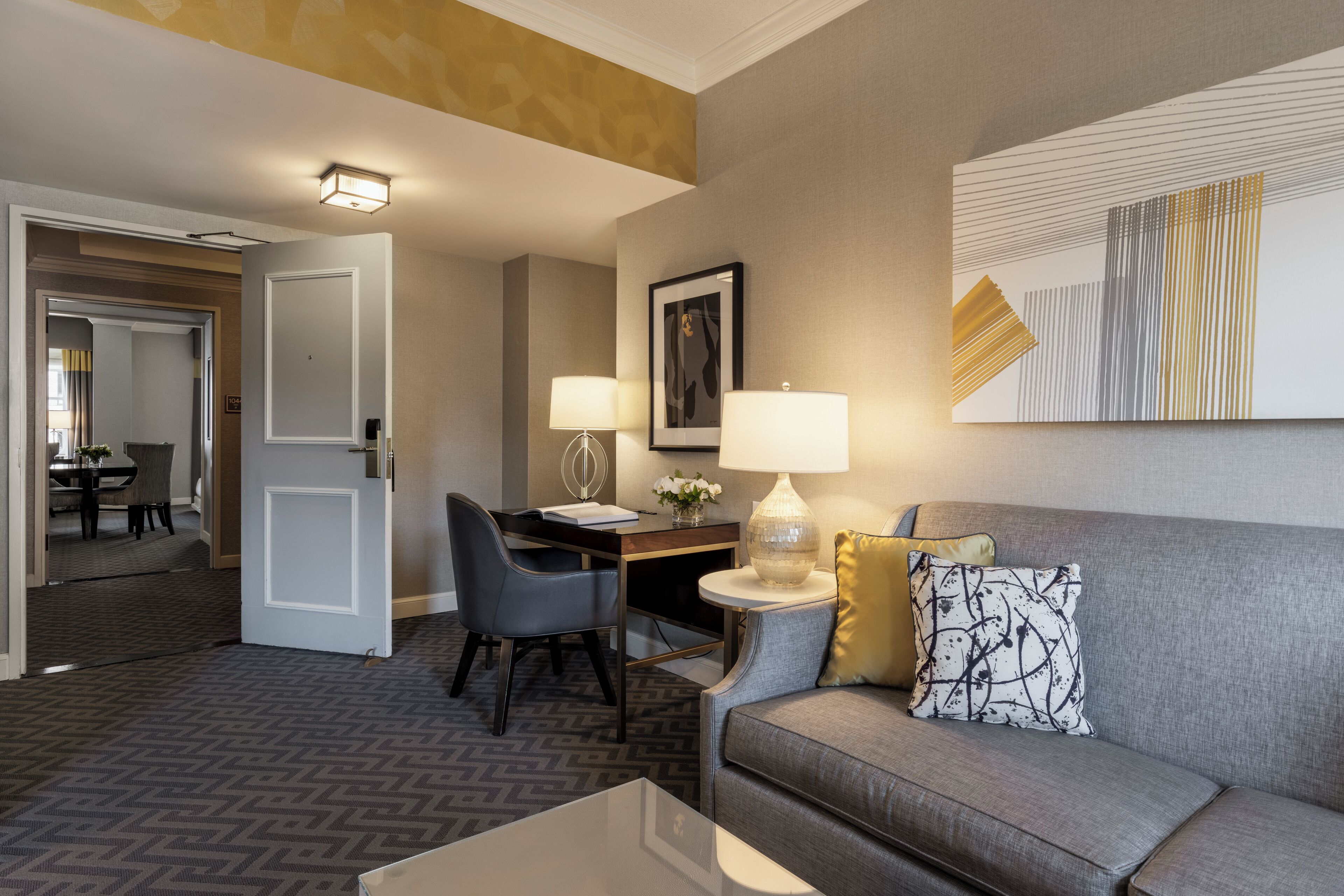 A hotel room with a gray sofa, a small table, a desk with two lamps, a framed picture on the wall, and a door leading to another room.