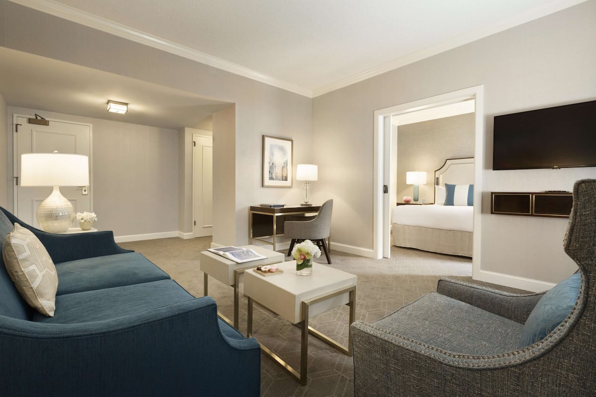 A modern hotel room with a blue sofa, a gray armchair, a small table with a lamp, a desk, and a flat-screen TV mounted on the wall.