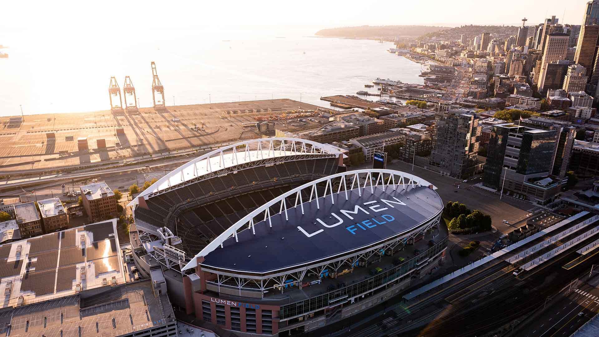 A stadium with "Lumen Field" written on the roof, surrounded by urban buildings and a waterfront.