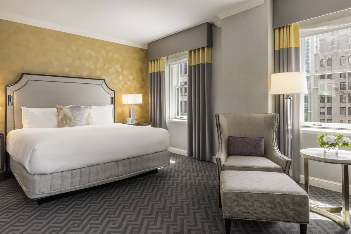 A hotel room with a large bed, a chair, a small table, and a window with curtains.