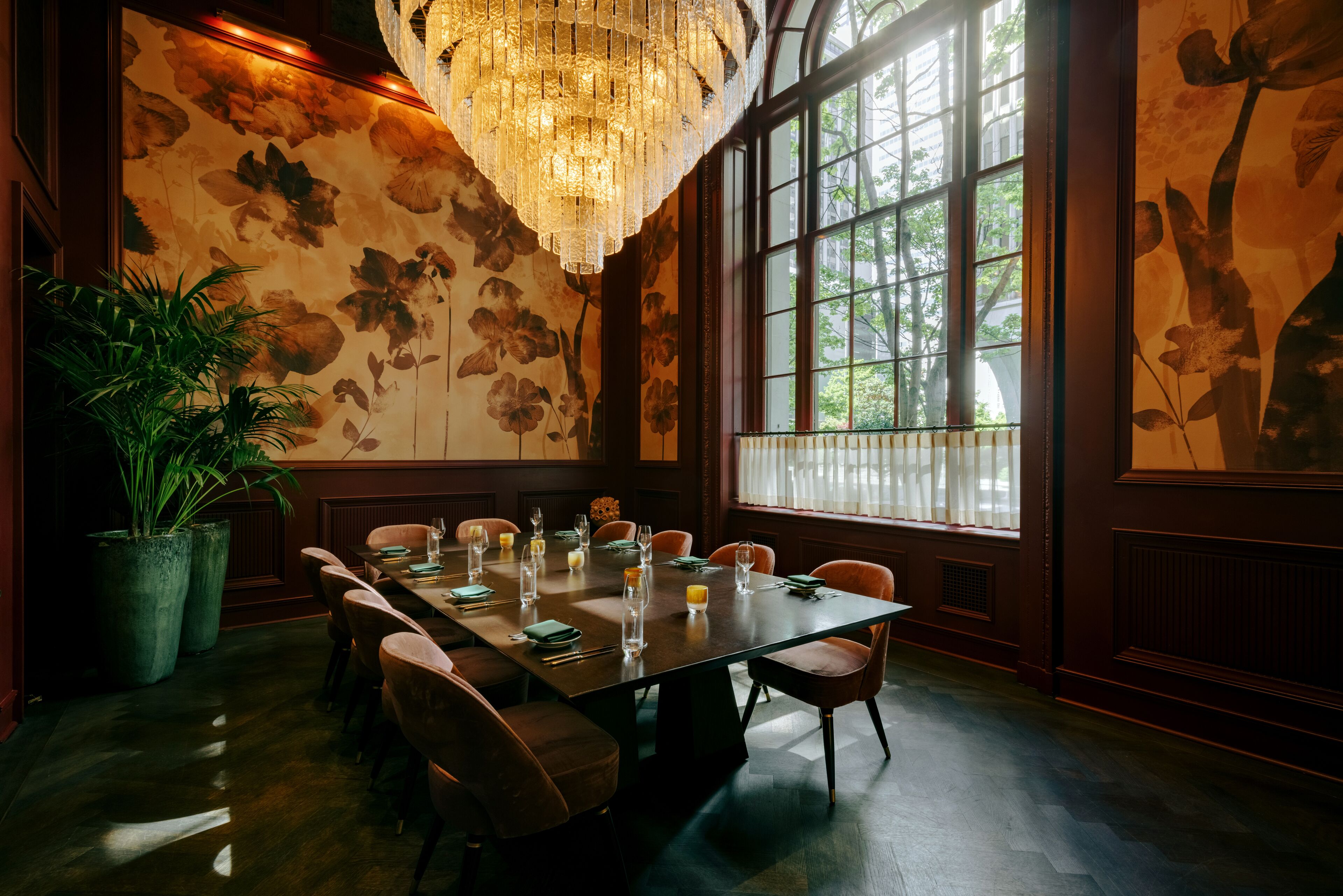 A dining room with a large table set for six, surrounded by chairs, with a chandelier above and a large window with a view of trees.