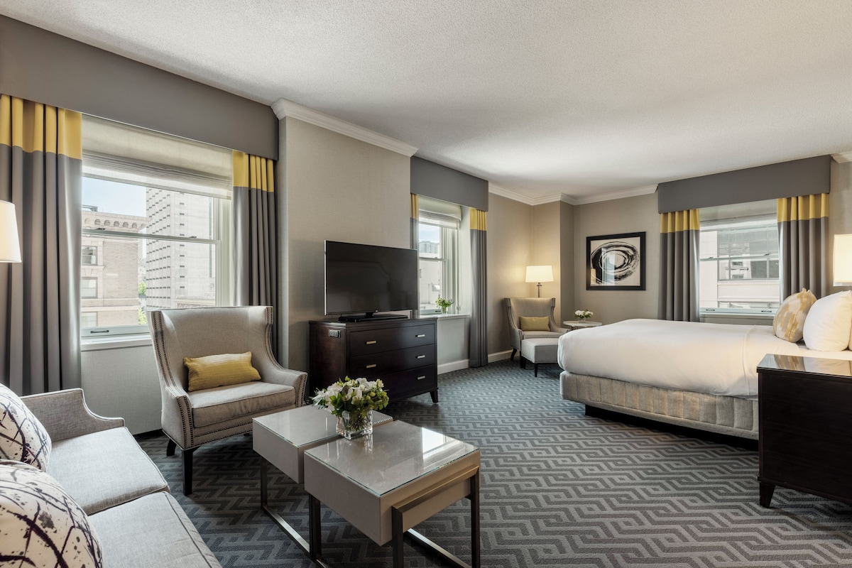 Une chambre d’hôtel avec un lit, un canapé, une chaise, une télévision, une commode et une fenêtre avec des rideaux.