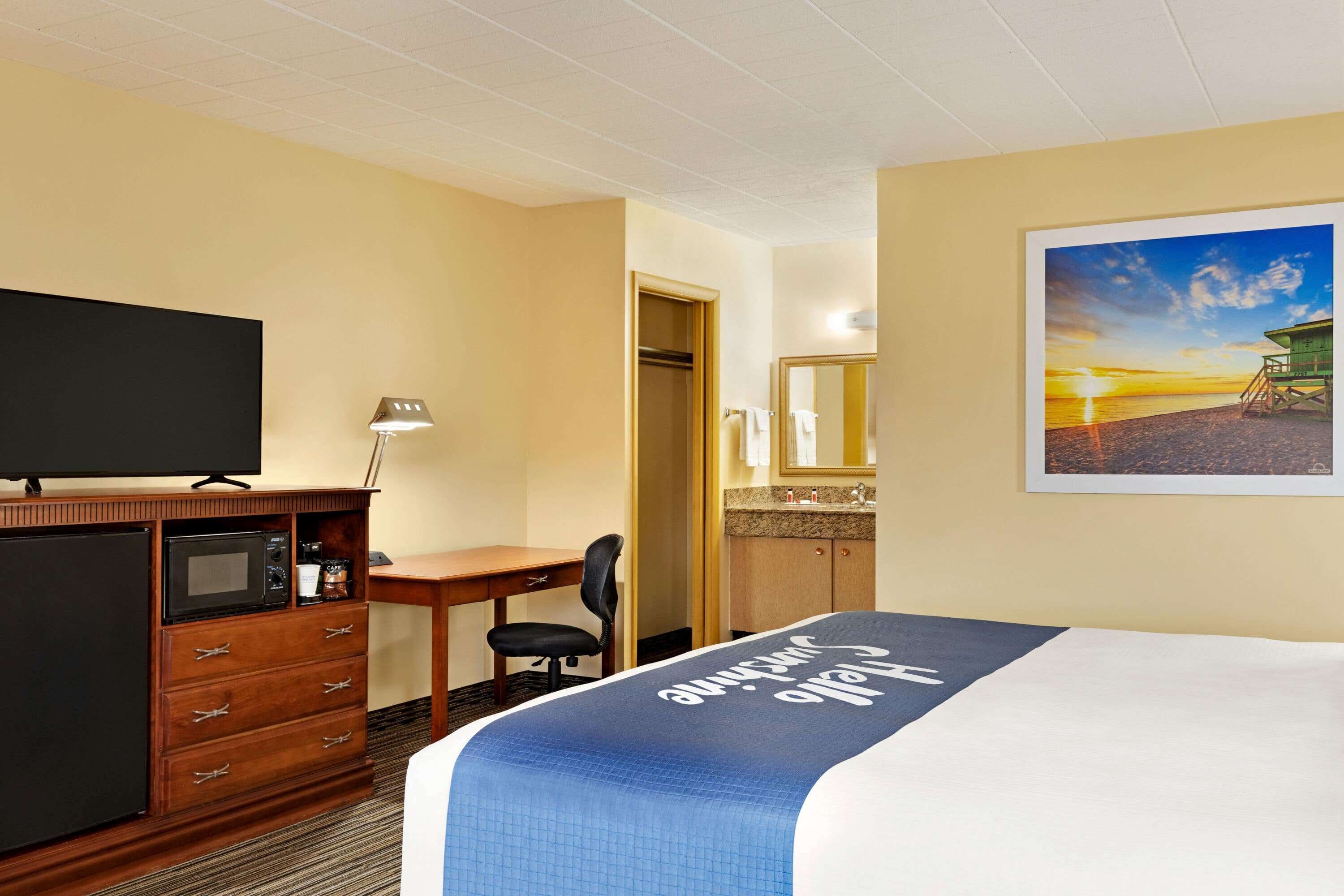 A hotel room with a bed, a desk, a television, a microwave, a coffee maker, and a bathroom with a sink and mirror.