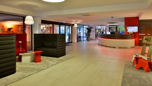 A modern hotel lobby with a reception desk, seating areas, and a large window offering a view of the outdoors. - Michel Hotel Wetzlar (Wetzlar)