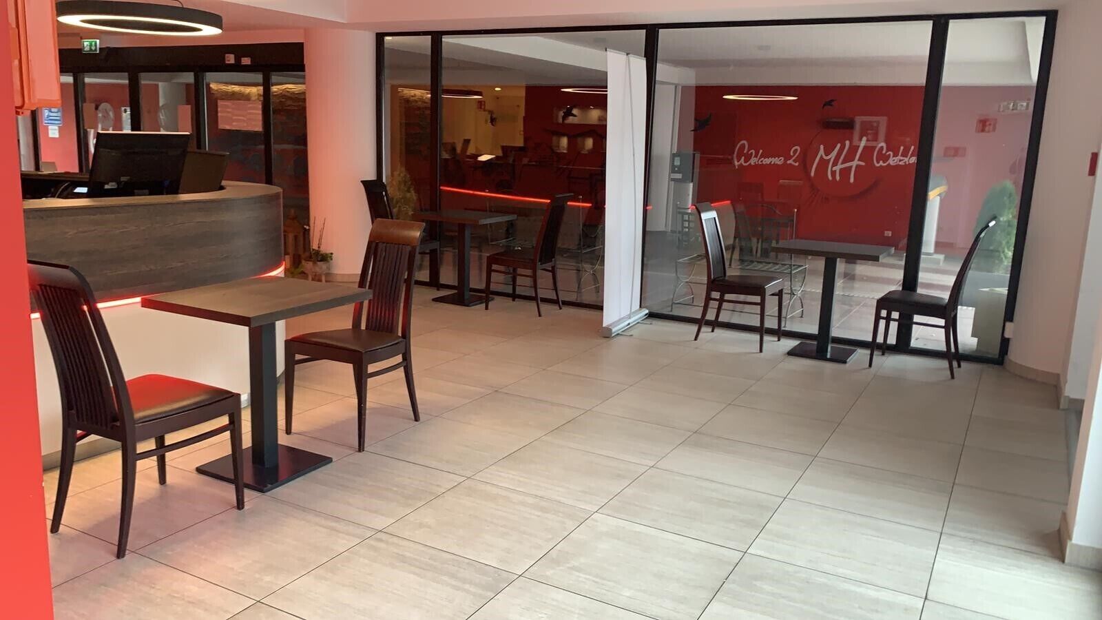 A modern dining area with wooden tables and chairs, a reception desk, and a glass wall with a red interior.