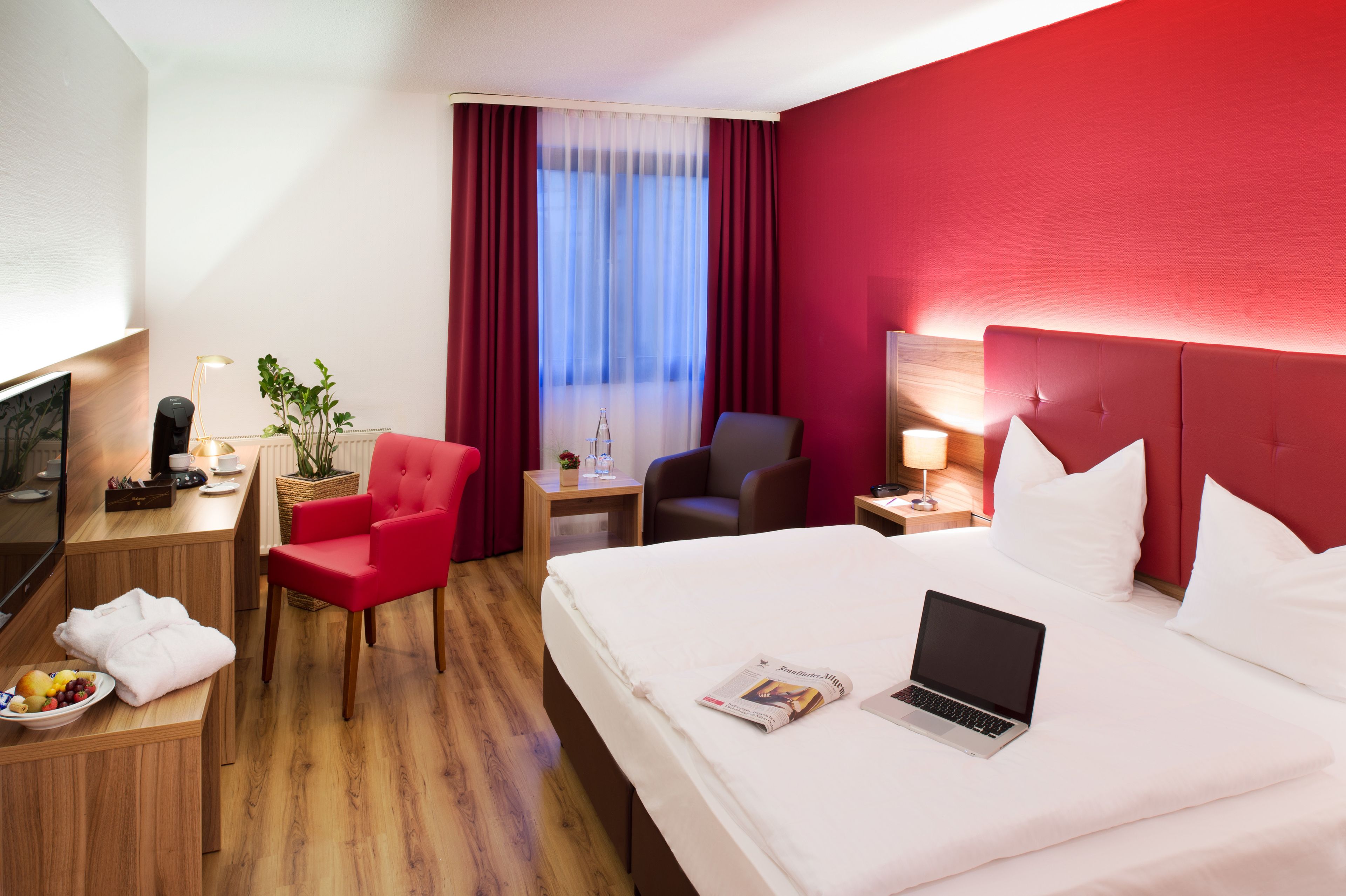 A hotel room with a bed, a desk with a laptop, a red chair, a small table with a plant, and a window with curtains.