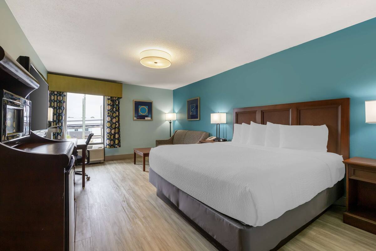 A hotel room with a large bed, a desk, a chair, a nightstand, and a window with curtains.