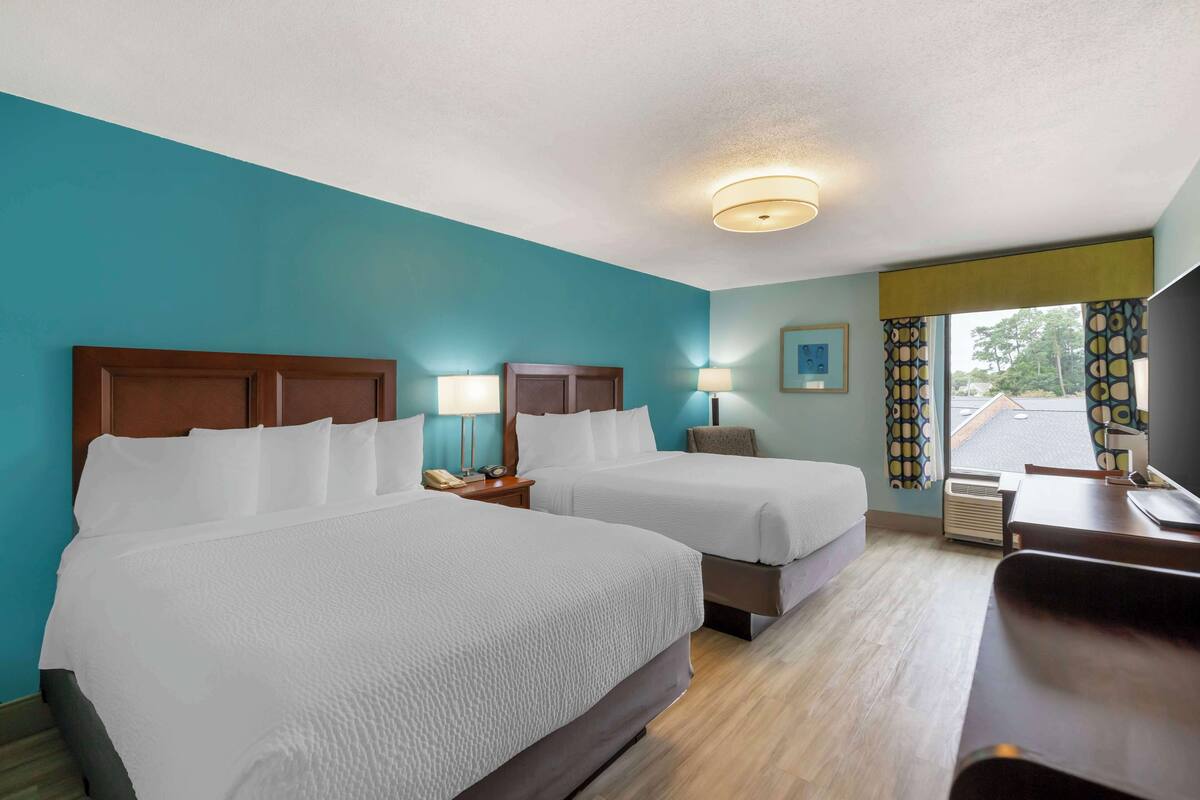 A hotel room with two beds, a desk, a chair, a lamp, and a window with curtains.