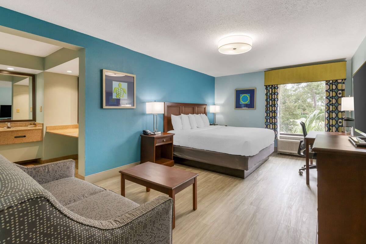 A hotel room with a bed, a desk, a chair, a nightstand, and a mirror.