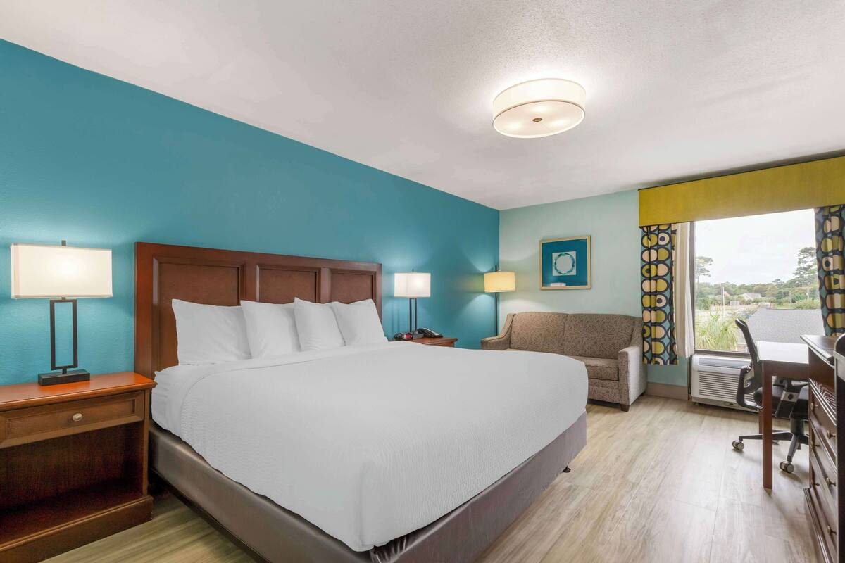 A hotel room with a large bed, two bedside tables with lamps, a desk with a chair, and a view of the outdoors through the window.