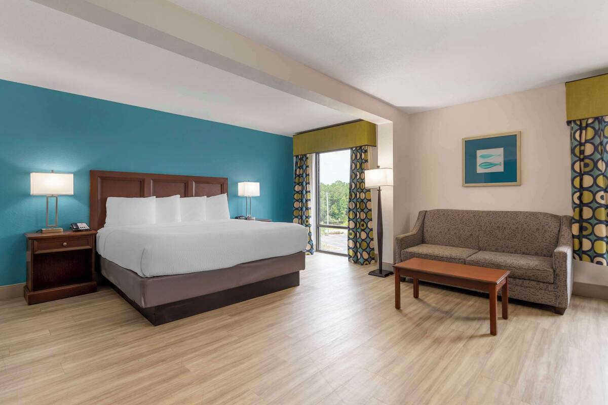 A hotel room with a large bed, a sofa, a nightstand with a lamp, and a window with curtains.
